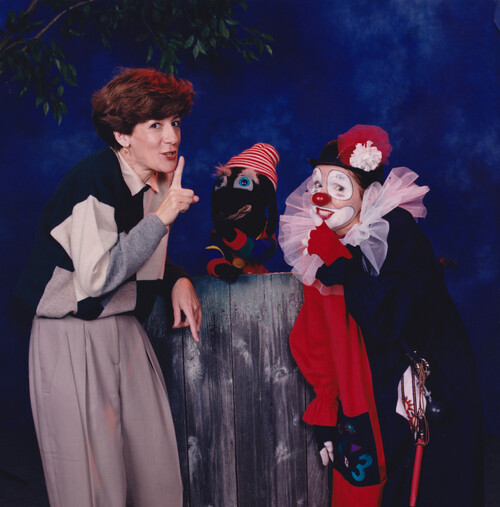 Suzanne Pinel, alias Marie-Soleil fait un geste de silence à côté d’une marionnette et d’un clown souriant, sur un fond de studio bleu.